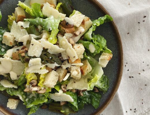 Caesars Salad mit Spargeln, gebratenen Pilzen, Brotwürfeln und hausgemachtem Dressing in einer blau-braunen Schüssel angerichtet.