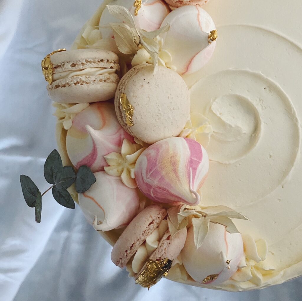 Von oben fotografierte Torte mit spiralförmiger Spachtelung, dekoriert mit Meringues und Macarons in rosa und beige von boumbelle